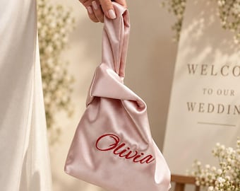 Puede incluir: Un bolso de nudo de satén rosa pálido con el nombre Olivia bordado en letra cursiva roja. El bolso tiene un acabado suave y brillante y un asa única formada por un diseño de tela anudada, sostenido por una mano con una pulsera de plata fina.