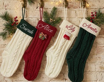 Puede incluir: Cuatro calcetines navideños de punto trenzado colgados de ganchos. El primero es crema con un puño verde oscuro bordado con Sarah. El segundo es rojo con un puño rojo bordado con Alice y un adorno de regalo. El tercero es crema con un puño crema bordado con Lily y un pequeño adorno de calcetín. El cuarto es verde oscuro con un puño crema bordado con Amelia.
