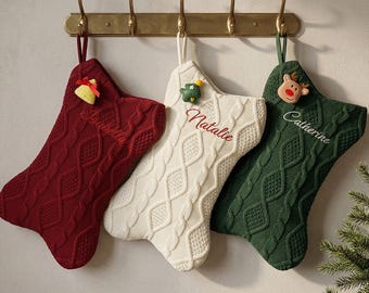 Puede incluir: Tres calcetines navideños de punto trenzado en color burdeos, crema y verde oscuro colgados de un perchero de latón. Cada calcetín tiene un nombre bordado en cursiva y un pequeño adorno de fieltro: una campana, un árbol y un reno.