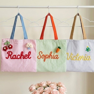 Può includere: Tre borse tote in velluto a coste appese a grucce bianche. La borsa rosa presenta il nome Rachel in rosso, un'applicazione a ciliegia e una nuvola. La borsa verde presenta il nome Sophia in arancione e una carota. La borsa viola presenta Victoria in giallo.