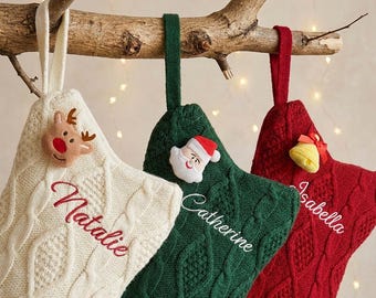 Puede incluir: Tres calcetines navideños de punto trenzado en color crema, verde oscuro y rojo. El calcetín crema tiene un reno, el verde una cara de Papá Noel y el rojo una campana amarilla. Cada calcetín está bordado con un nombre: Natalie, Catherine o Isabella.