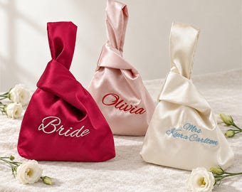 Puede incluir: Tres bolsos de nudo de satén en rojo oscuro, rosa claro y crema. El bolso rojo presenta el texto bordado en blanco "Bride", el rosa tiene el texto bordado en rojo "Olivia" y el crema tiene el texto bordado en azul "Mrs Kiara Carlton".