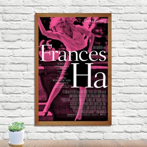 May include: A framed movie poster for "Frances Ha" featuring a pink-tinted image of a woman in motion. The title is prominently displayed in white, with the actress Greta Gerwig's name at the top. The poster includes quotes and credits.