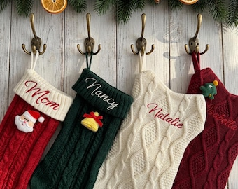 Puede incluir: Cuatro calcetines navideños de punto trenzado colgados en ganchos de metal. El calcetín rojo tiene una cara de Santa, el verde una campana amarilla, el blanco un patrón de trenzas y el rojo oscuro un pequeño árbol verde. Cada uno tiene un nombre.