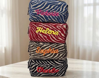 Puede incluir: Una pila de cuatro estuches rectangulares con cremallera y estampado de cebra. Cada estuche tiene nombres bordados en diferentes colores: azul con 'Victoria' rosa, rojo con 'Helena' amarillo, marrón con 'Sophia' naranja y negro con 'Charlie' rojo.