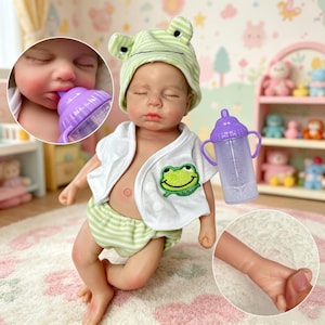 Könnte beinhalten: Eine lebensechte Babypuppe mit einem grün-weiß gestreiften Froschhütchen, passender Windel und einer weißen Jacke mit einem Froschaufnäher. Die Puppe hält eine lila Babyflasche. Nahaufnahmen zeigen das Gesicht und die Hand der Puppe.