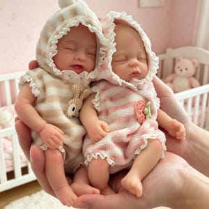 May include: Two realistic baby dolls, each wearing a hooded romper. One is beige striped with a bunny applique, the other is pink striped. Both dolls have closed eyes and are held in cupped hands.