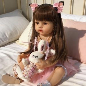 May include: A lifelike doll with long brown hair and bangs, wearing a pink dress and a headband with cat ears and pink bows. The doll holds a white stuffed bunny with floral print and the word "Love" on it. The doll is sitting on a bed.