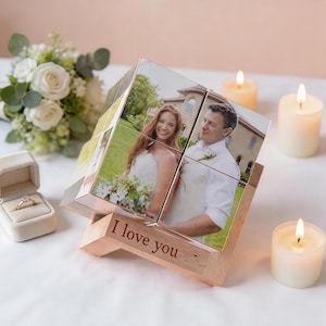 Könnte beinhalten: Ein personalisierter Fotowürfel mit Bildern eines Paares, auf einem Holzständer. Der Ständer ist mit den Worten "I love you" graviert. Der Würfel ist von Kerzen, einem Strauß weißer Rosen und einer Ringbox umgeben.