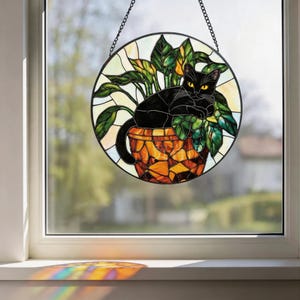 May include: Stained glass window art featuring a black cat lounging in a plant pot. The round window has a black metal frame and a chain for hanging. The cat has yellow eyes and is surrounded by green leaves and a mosaic-style pot.