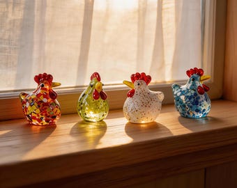 Puede incluir: Cuatro figuras de gallina de cristal con crestas rojas y picos amarillos se exhiben en un alféizar de madera. Las gallinas tienen diferentes patrones y colores, incluyendo un diseño de mosaico, un blanco moteado y un patrón floral azul. Las figuras están iluminadas por la luz del sol.