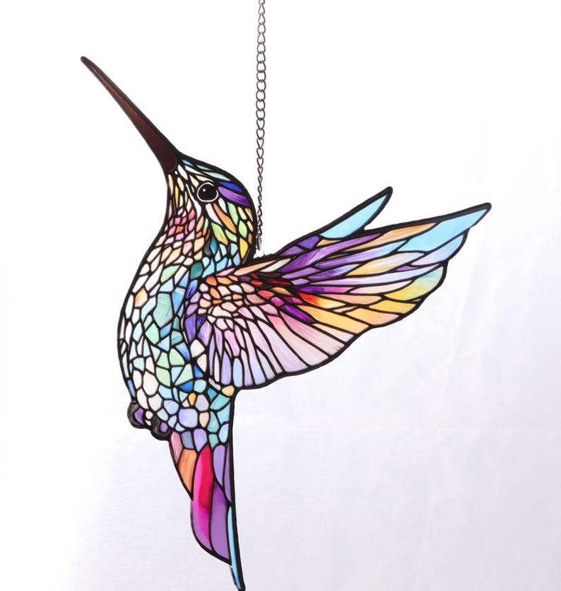 Puede incluir: Un colibr&iacute; de vidriera colorido con un pico largo y marr&oacute;n oscuro y alas extendidas. El cuerpo y las alas del ave est&aacute;n compuestos por varios colores, incluyendo azul, morado, amarillo y verde. Cuelga de una cadena de metal, perfecto para una ventana o pared.