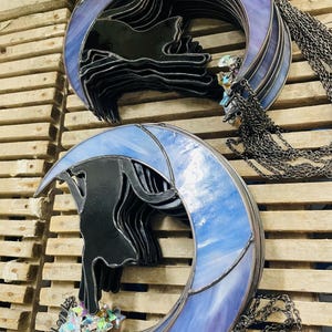 May include: Two stained glass suncatchers featuring a black cat silhouette perched on a crescent moon. One moon is purple, the other is light blue. Both include hanging metal chains and small iridescent star-shaped glass charms attached at the base.