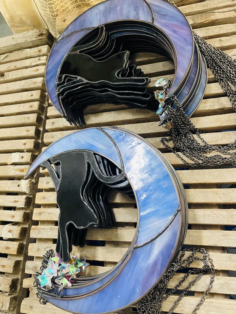 May include: Two stained glass suncatchers featuring a black cat silhouette perched on a crescent moon. One moon is purple, the other is light blue. Both include hanging metal chains and small iridescent star-shaped glass charms attached at the base.
