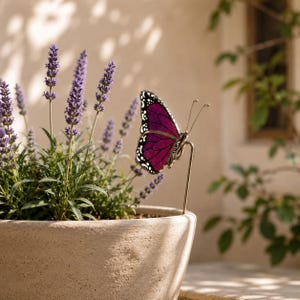 May include: A decorative butterfly with a deep magenta wing design and white and black accents, perched on a metal stake. The butterfly is next to a lavender plant in a beige pot. The scene is set outdoors with natural light.