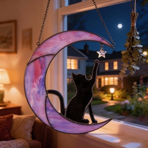 May include: A stained glass suncatcher shaped like a crescent moon in shades of pink and purple, featuring a black cat silhouette reaching towards a small, clear glass star charm. The piece hangs from a metal chain against a window background.