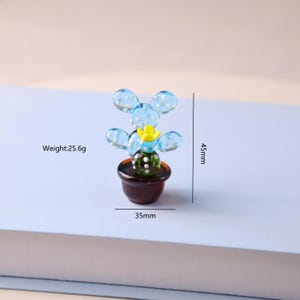 Puede incluir: Una escultura de cactus de vidrio en miniatura. El cactus presenta hojas redondeadas de color azul claro con lunares blancos, una flor amarilla y un centro verde oscuro. Est&aacute; en una maceta marr&oacute;n. La escultura mide 4,5 cm de alto y 3,5 cm de ancho, y pesa 25,6 g.