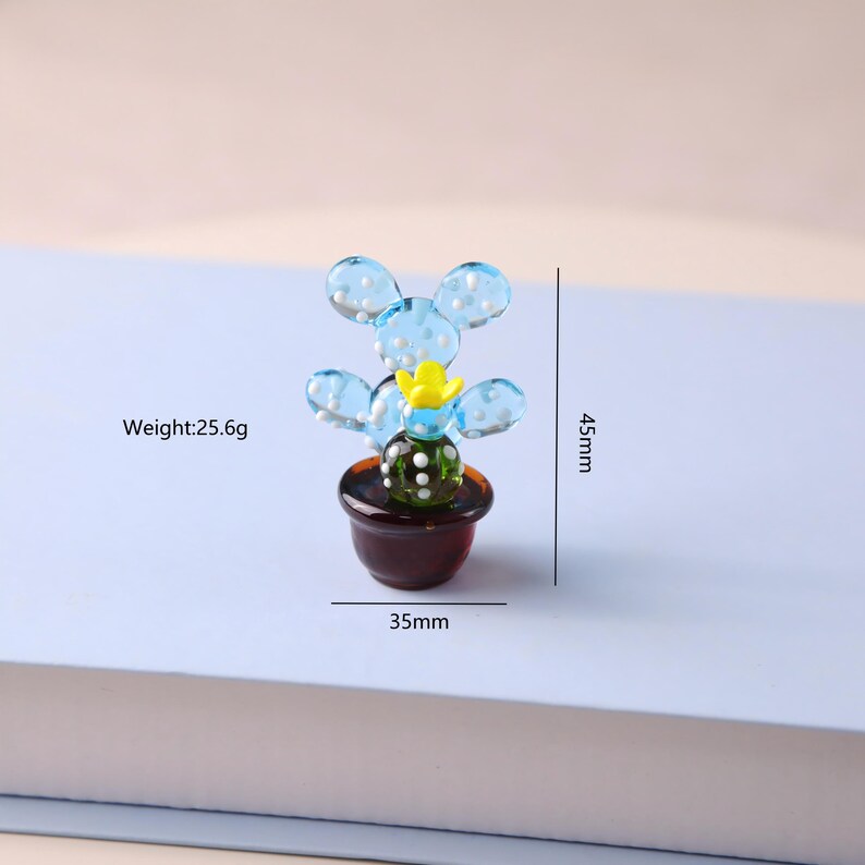 Puede incluir: Una escultura de cactus de vidrio en miniatura. El cactus presenta hojas redondeadas de color azul claro con lunares blancos, una flor amarilla y un centro verde oscuro. Est&aacute; en una maceta marr&oacute;n. La escultura mide 4,5 cm de alto y 3,5 cm de ancho, y pesa 25,6 g.