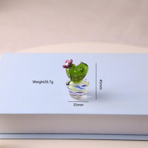 Puede incluir: Peque&ntilde;a escultura de cactus de vidrio con cuerpo verde en forma de coraz&oacute;n y flor rosa. El cactus est&aacute; en una maceta a rayas. La escultura mide 4,5 cm de alto, 3,5 cm de ancho y pesa 26,7 g. Un art&iacute;culo decorativo.