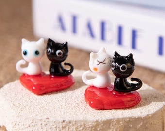 Puede incluir: Dos pares de figuras de gatos, cada uno sobre una base roja en forma de corazón. Un par presenta un gato blanco y un gato negro, el otro par un gato blanco y un gato negro. Las figuras están sobre una superficie texturizada blanquecina.