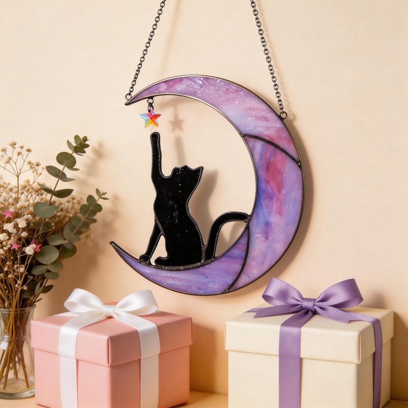 May include: A stained glass suncatcher featuring a crescent moon in shades of purple and pink with a black cat silhouette reaching towards a small, multi-coloured star charm. The piece hangs from a metal chain against a neutral background.
