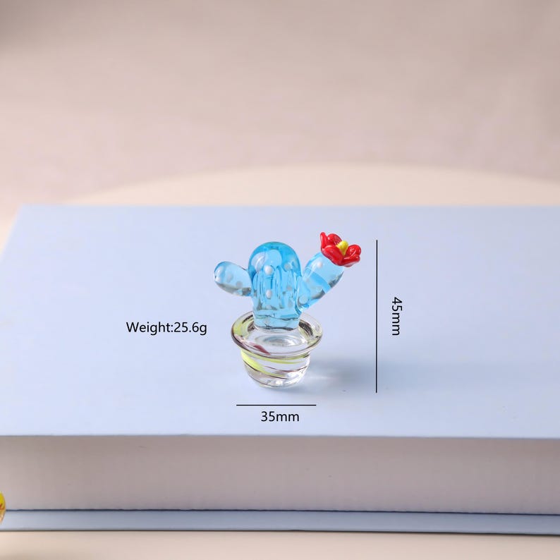 Puede incluir: Una peque&ntilde;a figura de cactus de cristal azul con una flor roja. El cactus est&aacute; en una maceta transparente con una base amarilla y verde. La figura mide 4,5 cm de alto y 3,5 cm de ancho y pesa 25,6 g.