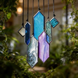 May include: A selection of stained glass pendants in shades of blue, purple, and green. Each pendant is framed in a dark metal and hangs from a black cord. The pendants are displayed against a backdrop of green foliage, creating a natural setting.