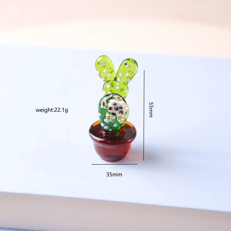 Puede incluir: Figura de cactus de cristal en una maceta roja. El cactus verde tiene lunares blancos y mide 5,5 cm de alto y 3,5 cm de ancho. La figura pesa 22,1 g. El cactus tiene dos estructuras verdes en forma de hoja en la parte superior.