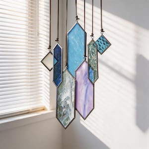 May include: A hanging mobile composed of seven geometric stained glass pieces in various shapes and sizes. The glass panels feature shades of blue, purple, white, and green with marbled and textured patterns, all framed in dark metal and suspended by cords.