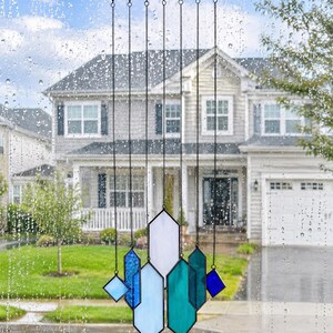 May include: A stained glass suncatcher featuring multiple geometric shapes in shades of blue, teal, and white, suspended by thin black metal chains from a horizontal black bar. The glass pieces are arranged in a vertical, layered composition.
