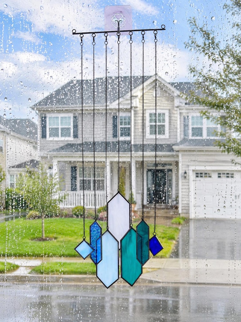 May include: A stained glass suncatcher featuring multiple geometric shapes in shades of blue, teal, and white, suspended by thin black metal chains from a horizontal black bar. The glass pieces are arranged in a vertical, layered composition.