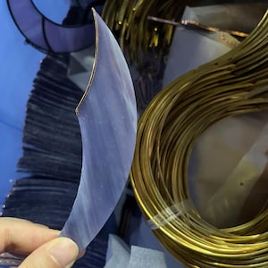 May include: A crescent-shaped piece of translucent purple stained glass with a copper foil edge. The glass features subtle white streaks and a smooth surface. Beside it sits a large coil of thin, flexible gold-coloured metal wire.