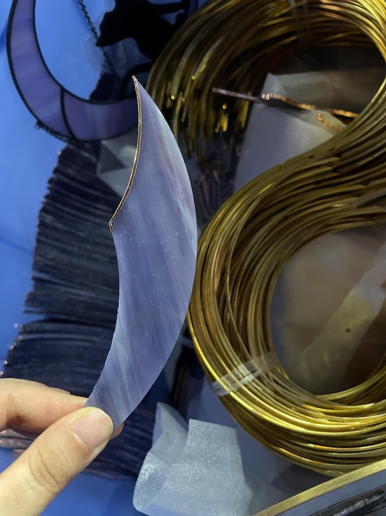 May include: A crescent-shaped piece of translucent purple stained glass with a copper foil edge. The glass features subtle white streaks and a smooth surface. Beside it sits a large coil of thin, flexible gold-coloured metal wire.