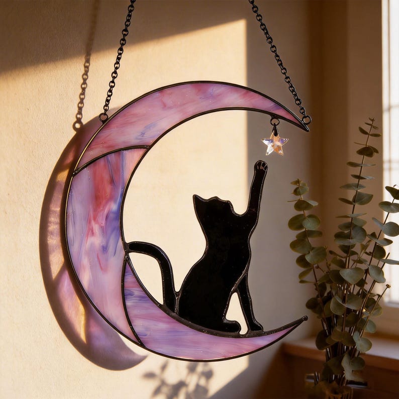 May include: A stained glass suncatcher featuring a crescent moon in shades of pink and purple with a black cat silhouette sitting on the moon, reaching up towards a small, clear, iridescent star charm suspended from the top of the moon frame.