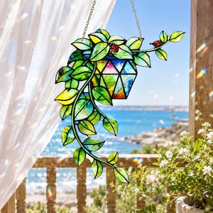 May include: A stained glass hanging plant decoration featuring a geometric pot filled with colorful glass. Green leaves and red ladybugs adorn the plant, which hangs from a chain. The background shows a view of the ocean.