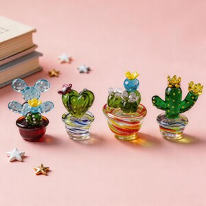 Puede incluir: Cuatro esculturas de cactus de vidrio de colores. Cada cactus tiene un dise&ntilde;o diferente, incluyendo un coraz&oacute;n y una corona. Los cactus est&aacute;n en peque&ntilde;as macetas a rayas. El fondo es rosa suave, con peque&ntilde;as decoraciones en forma de estrella.