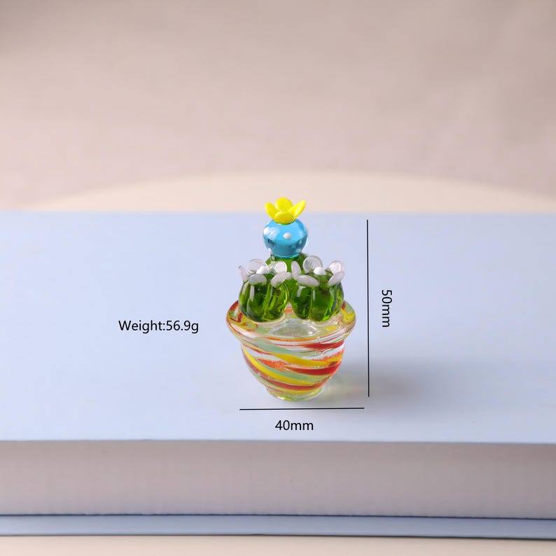 Puede incluir: Figura decorativa de cactus de cristal. El cactus es verde con una flor amarilla y detalles azules. La base es una maceta a rayas con rojo, amarillo y naranja. La figura mide 5 cm de alto y 4 cm de ancho. Peso: 56,9 gramos.