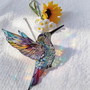 Puede incluir: Un atrapasoles de colibr&iacute; colorido con un dise&ntilde;o de mosaico. El p&aacute;jaro tiene un pico largo y negro y una cadena plateada para colgarlo. Las alas son en tonos de amarillo, naranja, morado y azul. Un girasol y margaritas est&aacute;n en el fondo.