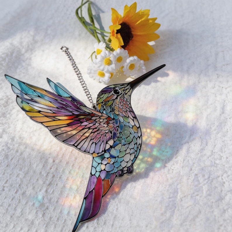 Puede incluir: Un atrapasoles de colibr&iacute; colorido con un dise&ntilde;o de mosaico. El p&aacute;jaro tiene un pico largo y negro y una cadena plateada para colgarlo. Las alas son en tonos de amarillo, naranja, morado y azul. Un girasol y margaritas est&aacute;n en el fondo.