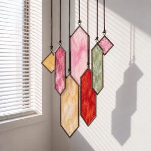 May include: A hanging mobile featuring multiple geometric stained glass panels in shades of pink, red, yellow, and green. Each panel is framed in dark metal and suspended by individual brown cords, creating a layered, vertical arrangement.