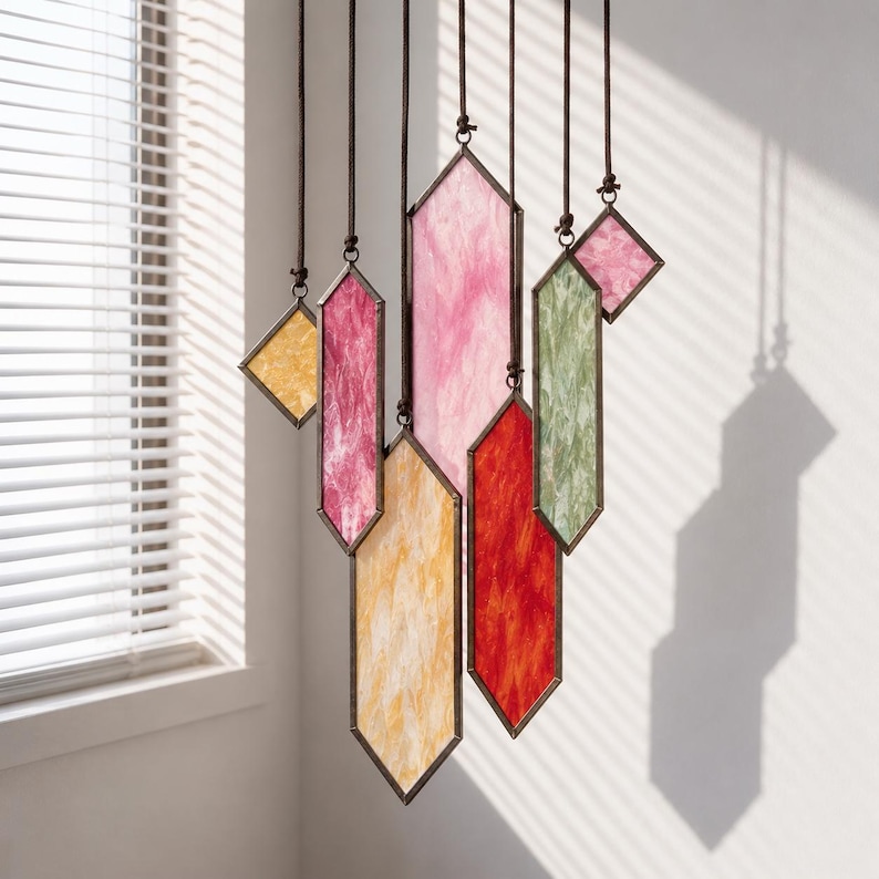 May include: A hanging mobile featuring multiple geometric stained glass panels in shades of pink, red, yellow, and green. Each panel is framed in dark metal and suspended by individual brown cords, creating a layered, vertical arrangement.