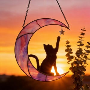 May include: A crescent moon suncatcher made of pink and purple marbled stained glass with a black metal frame. A black cat silhouette sits on the moon, reaching up towards a small, clear, five-pointed star charm hanging from the top curve.