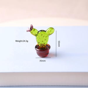 Puede incluir: Peque&ntilde;a figura decorativa de cactus de vidrio. El cactus es verde con una superficie texturizada y una peque&ntilde;a flor rosa. Est&aacute; en una maceta roja. La figura mide 5,5 cm de alto, 3,5 cm de ancho y pesa 24,3 g.