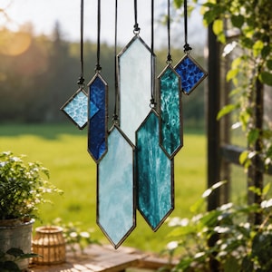 May include: A hanging mobile featuring multiple geometric stained glass pieces in shades of blue, teal, and white. The glass shapes include diamonds and elongated hexagons, each framed in dark metal and suspended by thin black cords at varying heights.