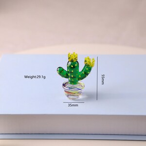 Puede incluir: Una figura de cactus de vidrio verde con detalles amarillos en forma de corona. El cactus est&aacute; en una maceta de vidrio colorida. La figura mide 5,5 cm de alto y 3,5 cm de ancho. El peso es de 29,1 g.