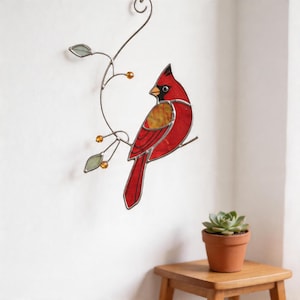 Puede incluir: Un adorno de pared de un pájaro cardenal de vidrieras. El pájaro es principalmente rojo con detalles en negro y está posado en una rama de metal con hojas y cuentas naranjas. El pájaro está montado en una pared blanca.