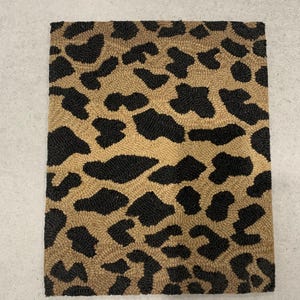 May include: A rectangular rug featuring a dense, tufted texture with a repeating leopard print pattern. The design consists of irregular black spots set against a tan, beige-colored background, creating a high-contrast, textured surface.
