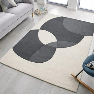 May include: A contemporary rectangular rug with a geometric design. The rug has a cream-coloured base with overlapping grey shapes. It's placed on a light wooden floor, with a sofa and chair in the background. The rug's design adds a modern touch to the room.