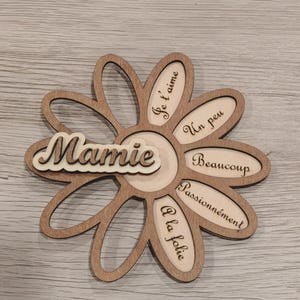 Peut inclure: Une décoration en forme de fleur en bois avec le mot "Mamie" en écriture cursive. Les pétales de la fleur portent des phrases telles que "Je t'aime" et "Passionnément". La décoration est en bois brun clair.