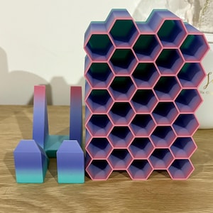 Puede incluir: Un conjunto de objetos geométricos multicolores. La pieza principal es una estructura en forma de panal con tonos rosas, azules y verdes. Otras piezas incluyen pequeñas casas y rectángulos, todos con un degradado de color. Los objetos están sobre una superficie de madera.